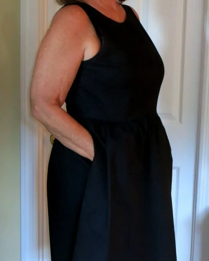 Femme aux cheveux châtains, en robe noire ajustée, debout sur un balcon avec une porte ouverte derrière elle.