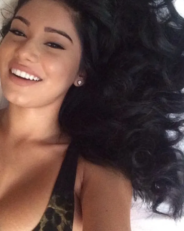 Femme allongée, cheveux noirs bouclés, souriant, sur un lit blanc.